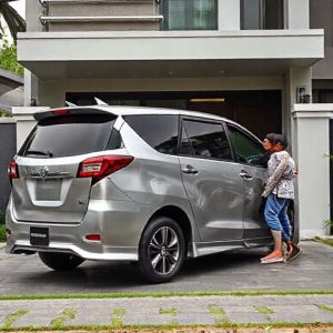 Toyota Innova Reborn warna silver diparkir di depan rumah modern, dengan keluarga kecil (ayah, ibu, 2 anak) sedang menaruh barang ke bagasi. Latar belakang pepohonan hijau.