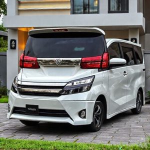 Toyota Voxy bekas berwarna putih sedang parkir di depan rumah modern, menunjukkan pintu geser terbuka dan keluarga muda tersenyum di dekatnya. Langit cerah, ada sentuhan hijau dari tanaman.